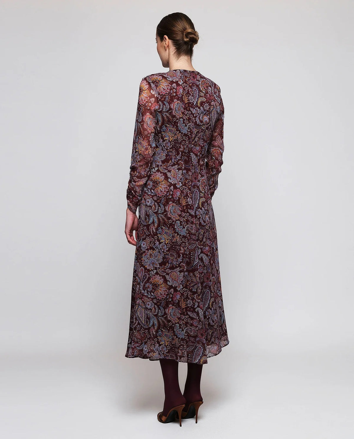 A woman stands facing away, wearing the MIRTO 1956 Burgundy Draped Paisley Print Dress—a long sleeve midi dress featuring an elegant paisley pattern—paired with burgundy tights and black heels. Her hair is styled in a neat bun as she poses against a plain white background.