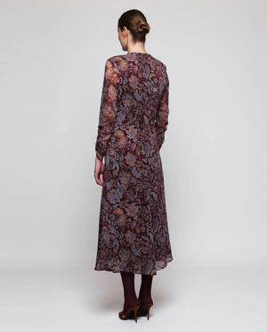 A woman stands facing away, wearing the MIRTO 1956 Burgundy Draped Paisley Print Dress—a long sleeve midi dress featuring an elegant paisley pattern—paired with burgundy tights and black heels. Her hair is styled in a neat bun as she poses against a plain white background.