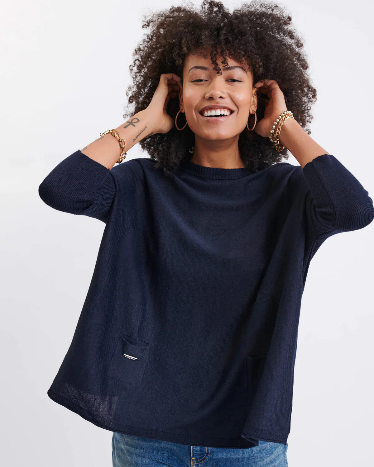 A smiling woman with curly hair wears the MERSEA - CATALINA CREWNECK SWEATER in navy blue and jeans, standing against a plain light background with her hands near her head.