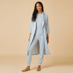 A woman wearing the SOFTIES Dream Shawl Collar Robe in Sky Blue Stripe by SOFTIES sits on a wooden chair, adjusting her hair with one hand and resting her other arm on the chair’s back against a beige background, evoking spa-like luxury akin to slipping into this lightweight robe.