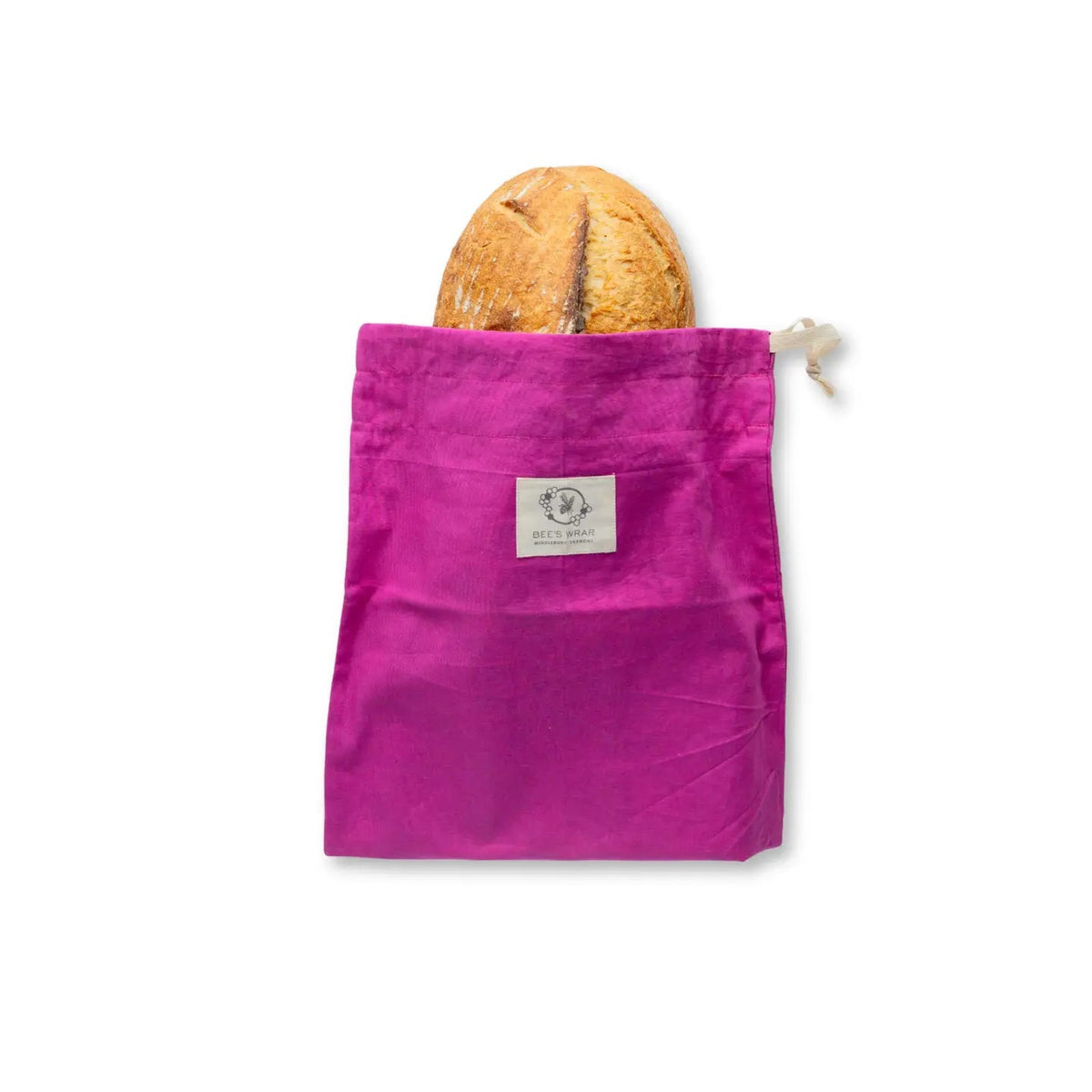 A round loaf of bread in the BEES WRAP - BEESWAX REUSABLE BREAD BAKERY BAG MAGENTA by BEES WRAP, featuring a magenta cloth and floral trim, is placed on a textured tablecloth beside a wooden cutting board, wheat stalks, and a folded napkin.