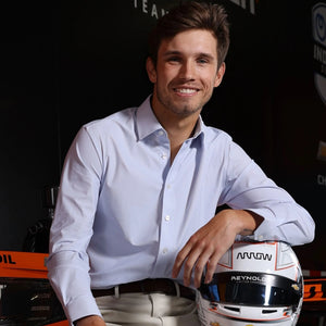 A man in a COLLARS AND CO Semi Spread Collar Apex Performance Dress Shirt in White and Blue Cornerstone smiles beside a racing helmet, his arm resting on it, with racing-themed decor and logos in the background.
