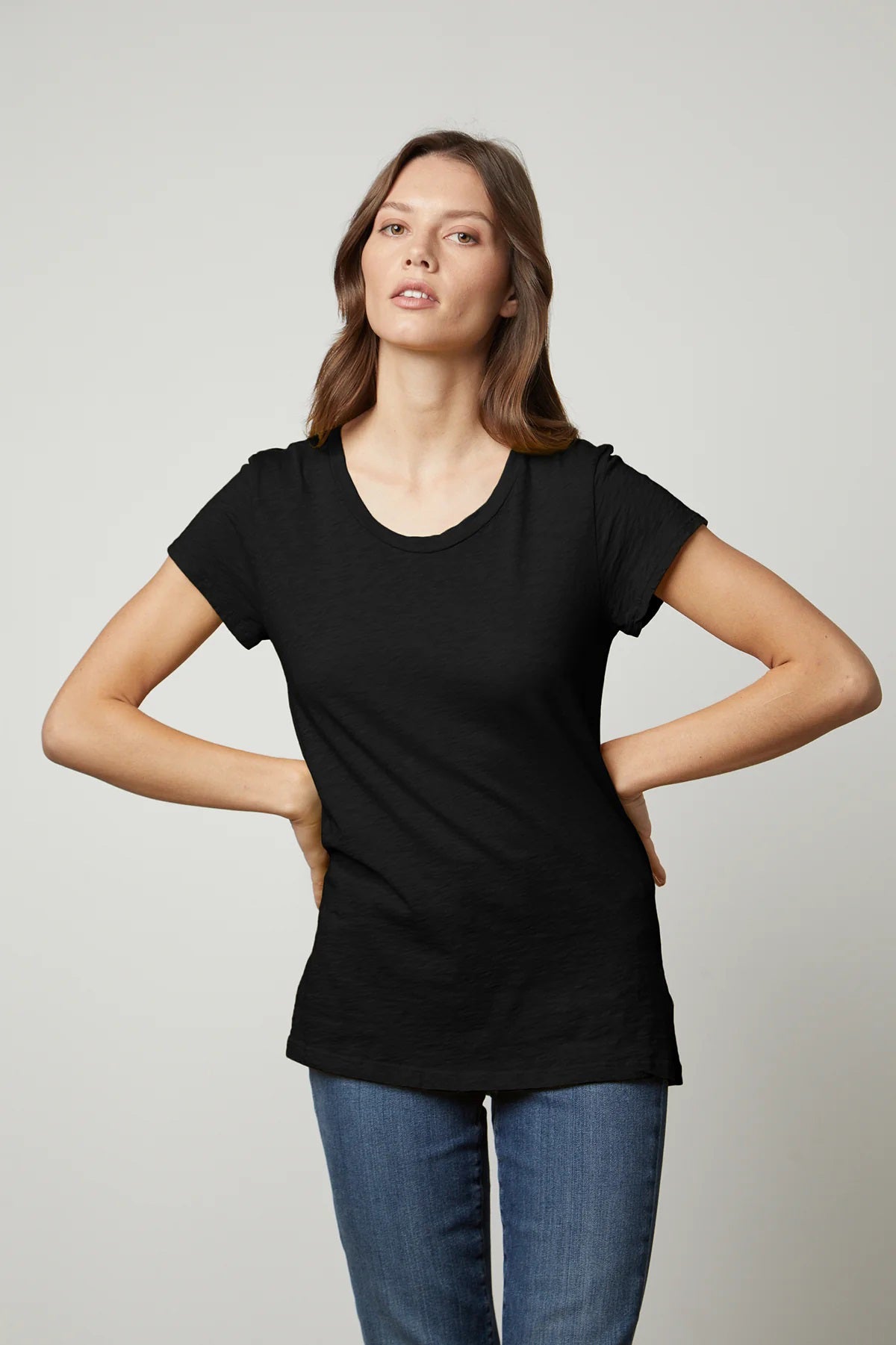 A person with long dark hair is standing against a neutral background, wearing the VELVET - ODELIA CREW NECK COTTON SLUB TEE from VELVET BY GRAHAM &amp; SPENCER and blue jeans. Their hands are resting on their hips as they look straight ahead with a neutral expression.