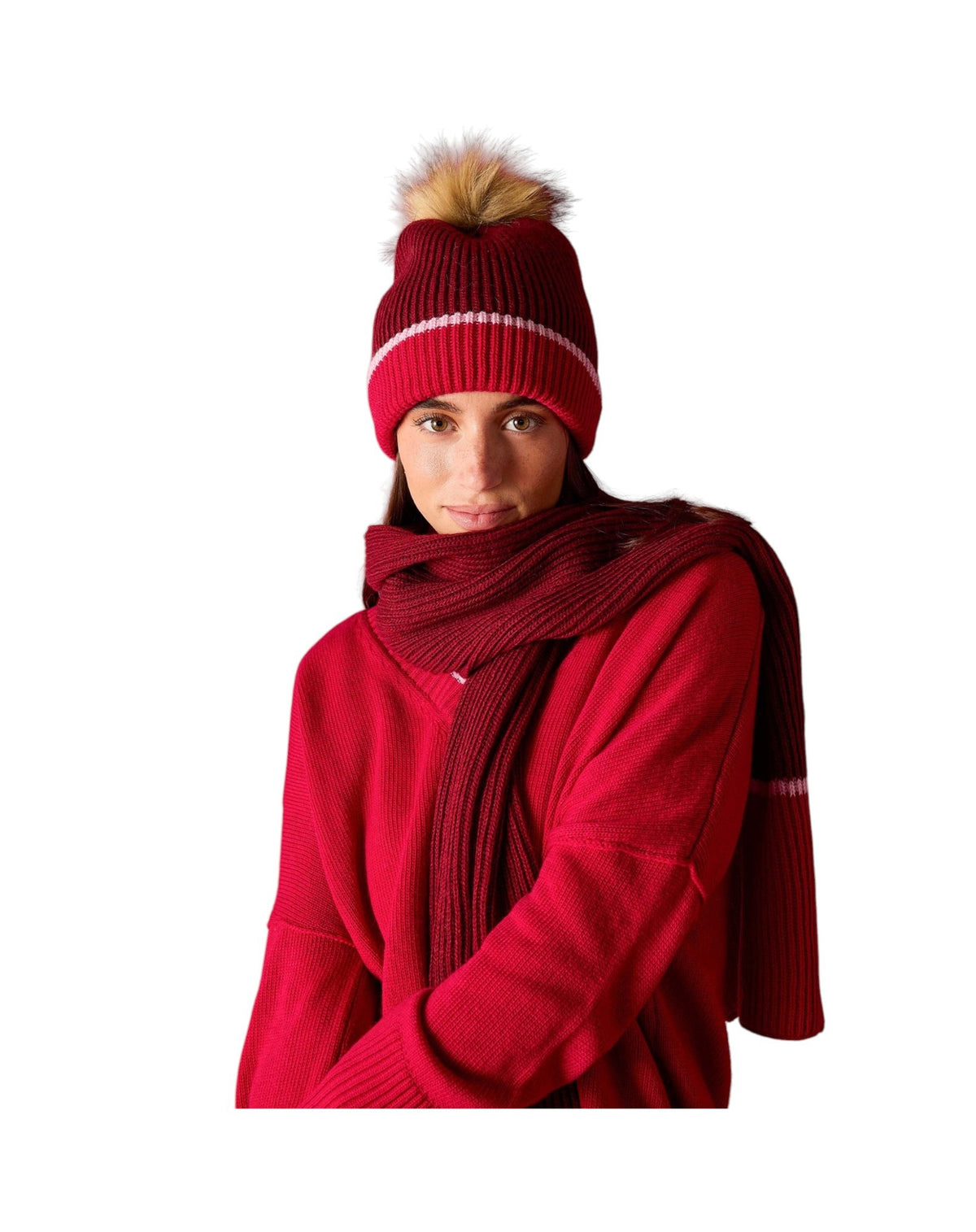 The MERSEA - Montauk Colorblock Beanie in Raspberry by MERSEA is a chunky knit red beanie with a folded brim, featuring a white stripe above the brim and finished with a light brown faux fur pom-pom, shown against a white background.