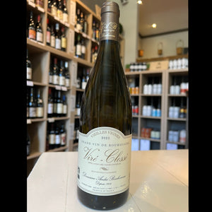 A bottle of BONHOMME VIRÉ-CLESSÉ 'VIELLES VIGNES' 2023 by WINE: PROOF sits on a white table in a wine shop, surrounded by shelves filled with assorted bottles and jars in the background.