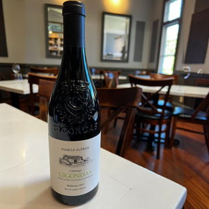 A bottle of FAMILLE AUTRAN GIGONDAS 2021 from WINE: PROOF rests on a white table in a restaurant, surrounded by wooden chairs, tables, and large windows in the background, embodying the elegance of Southern Rhône.