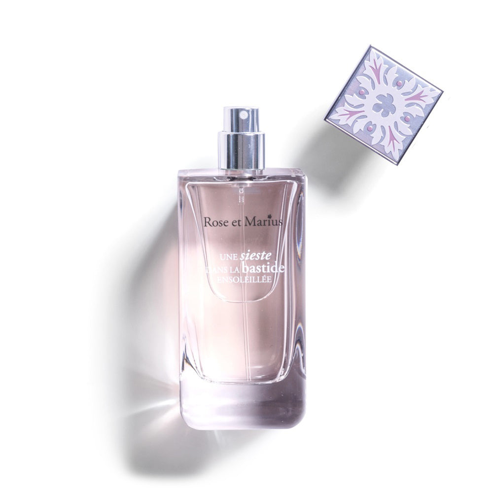 A clear ROSE ET MARIUS - A SIESTA IN A SUNLIT HOME EAU DE PARFUME 100ML bottle with a silver spray top and patterned square cap evokes summer in Provence, embodying the floral spirit of the ROSE ET MARIUS brand on a white background.