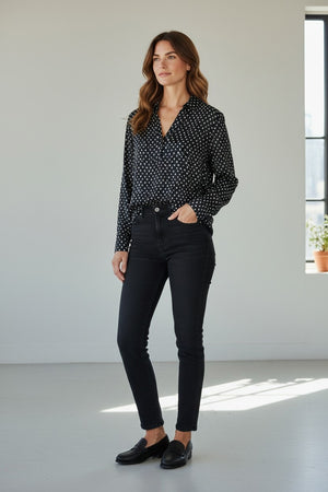 A woman stands in a bright room wearing the HARTFORD - CHORUS POPOVER SHIRT IN NAVY WITH WHITE DOTS, crafted by HARTFORD from soft printed viscose. She pairs it with black jeans and classic black loafers, one hand in her pocket as she gazes slightly to the side, radiating timeless French elegance. A small plant sits on a windowsill in the background.