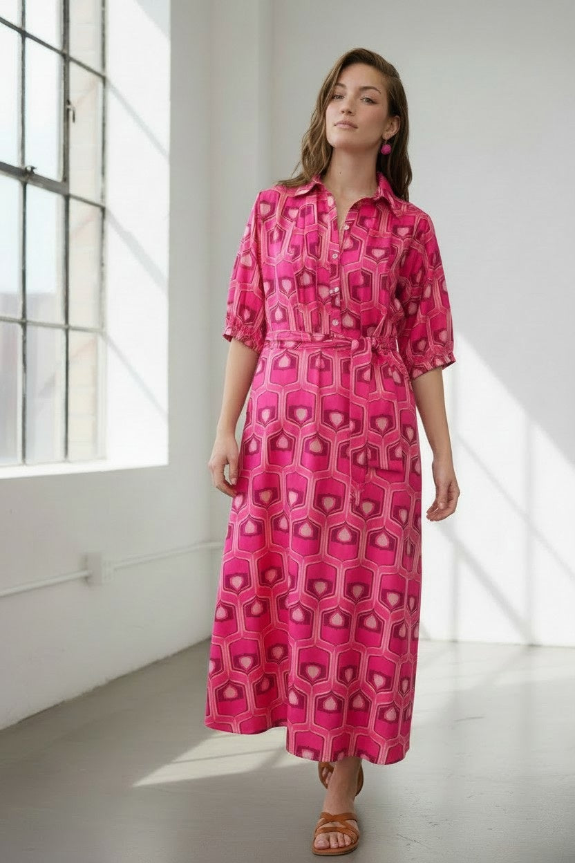 A woman stands indoors wearing the FLOOR Cotton Maxi Dress in a bright pink geometric print, featuring short sleeves. Sunlight streams through large windows, illuminating the minimalist room as she pairs the look with sandals.