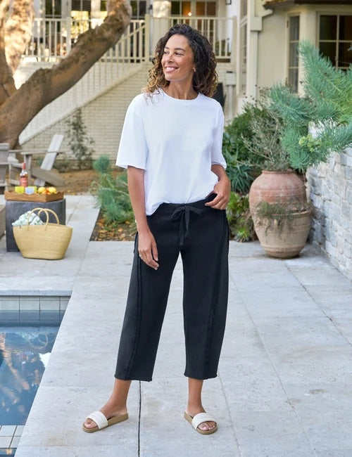A person wearing the FRANK & EILEEN Bibi Barrel Leg Sweatpant in Triple Fleece British Royal Navy, styled with a drawstring waist and barrel-leg silhouette, along with white slide sandals, stands on a stone patio near greenery and a large clay pot.