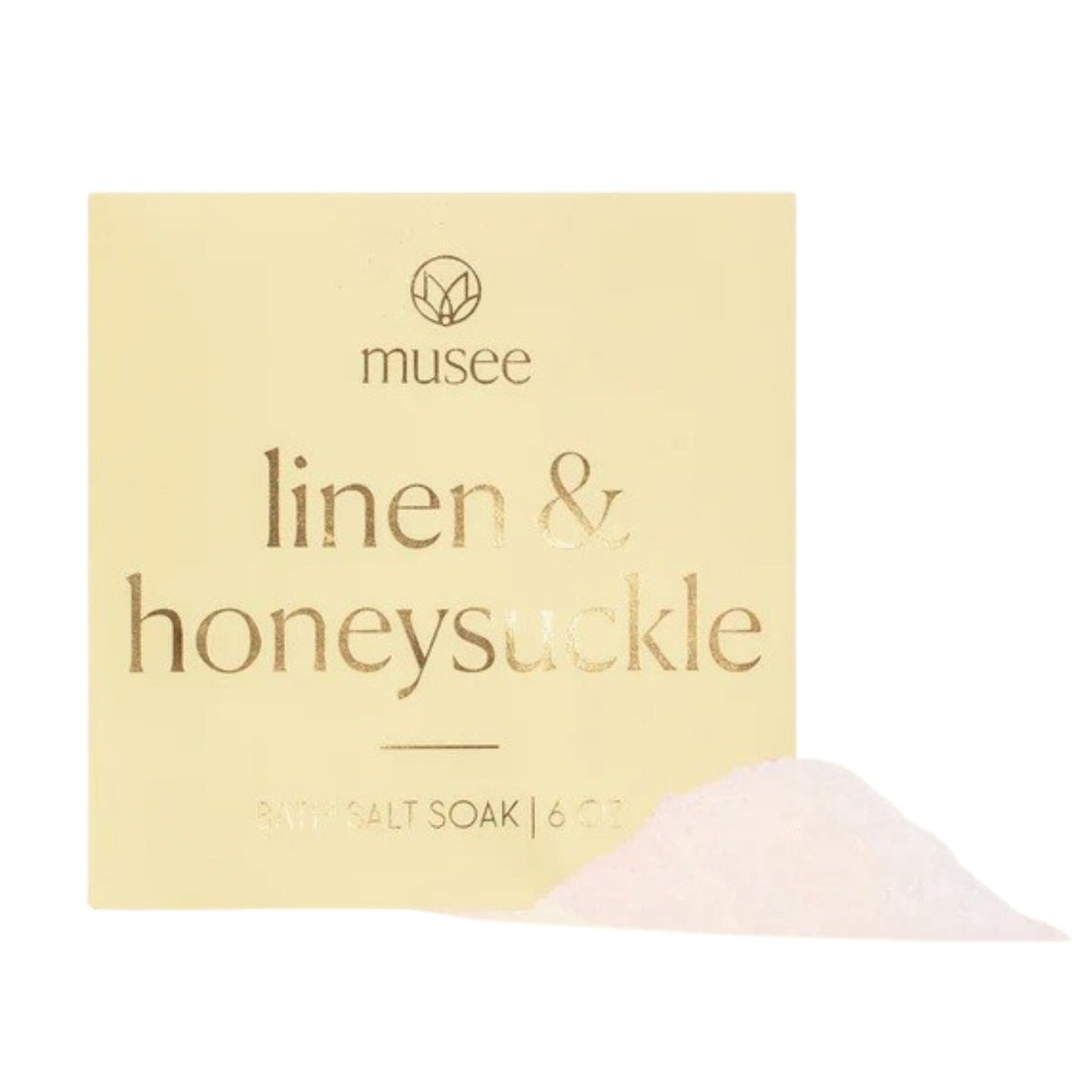 A package labeled “MUSEE - LINEN & HONESUCKLE MINI BATH SALT SOAK” from MUSEE rests on a white plate beside a green succulent on a wooden surface, highlighting exquisite natural bath products crafted with Pacific sea salt for the ultimate in relaxation.
