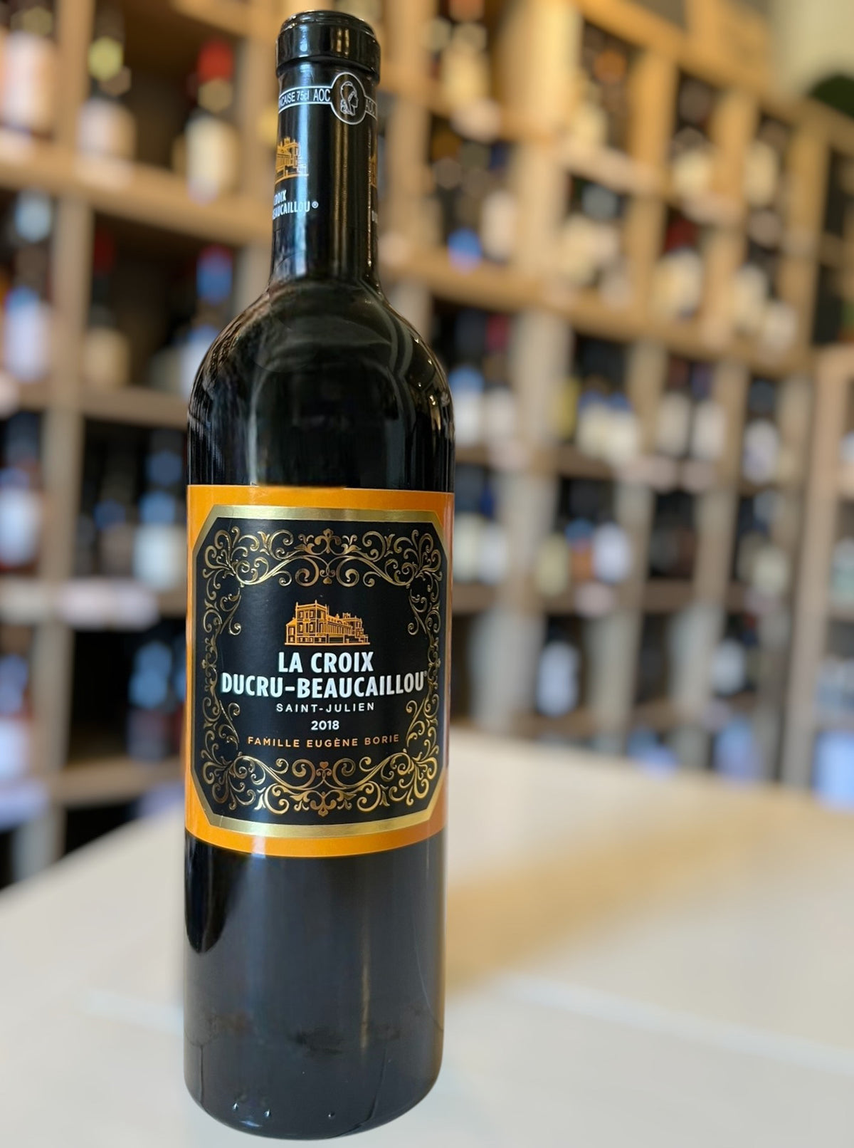 A bottle of CHÂTEAU DUCRU-BEAUCAILLOU ‘LA CROIX’ 2018 from WINE: MONSIEUR TOUTON, adorned with an ornate gold and black label, stands upright against a white background. This refined 2018 vintage highlights the esteemed character of the Saint-Julien appellation.