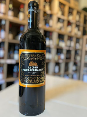 A bottle of CHÂTEAU DUCRU-BEAUCAILLOU ‘LA CROIX’ 2018 from WINE: MONSIEUR TOUTON, adorned with an ornate gold and black label, stands upright against a white background. This refined 2018 vintage highlights the esteemed character of the Saint-Julien appellation.