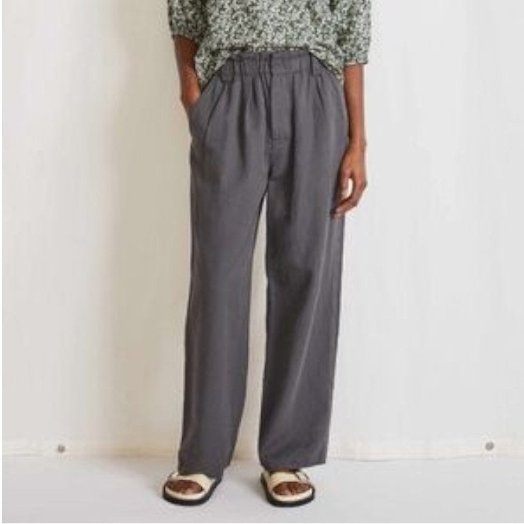 A person poses against a white background wearing a patterned top, the APIECE APART SON VIDA PANT in gray made from organic cotton, and sandals, with their hands resting comfortably in the pockets of the pants.