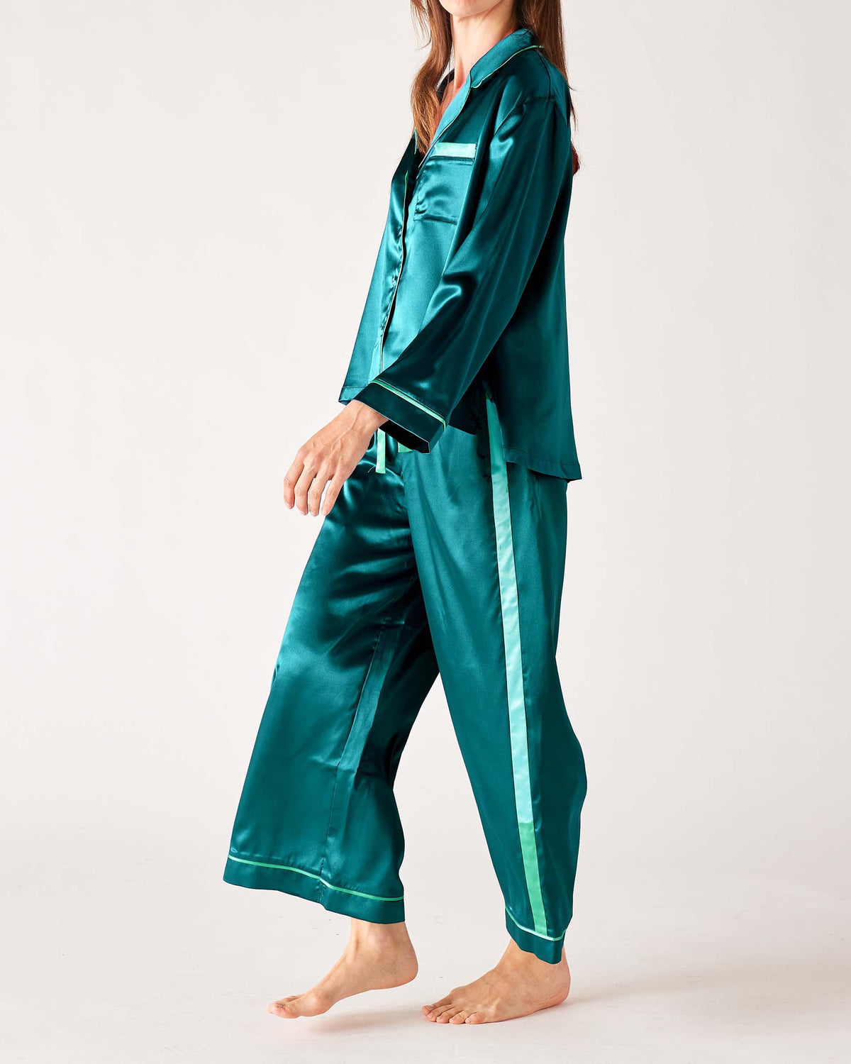 A woman models the MERSEA - OVER THE MOON SATIN SAILOR PAJAMA SET in teal with white trim, featuring a long-sleeved button-up top and elastic waist bottoms. She stands barefoot with her hands in the pockets, highlighting the silky smooth fabric.