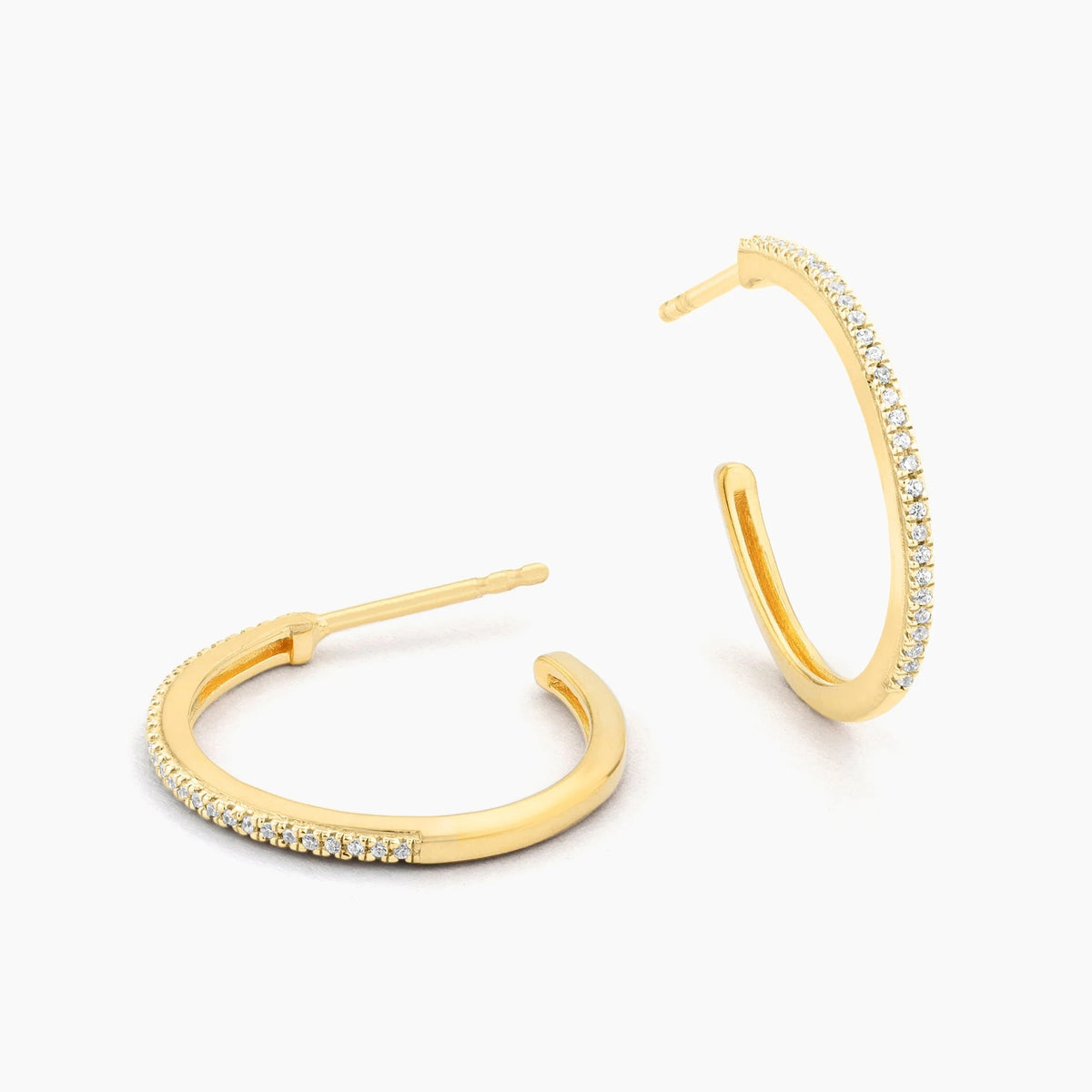 The ELLA STEIN - A HOOP THING EARRINGS by ELLA STEIN are gold-plated hoop earrings adorned with small clear stones along the outer edge, showcased on a plain white background.