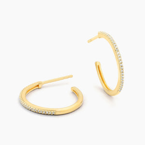 The ELLA STEIN - A HOOP THING EARRINGS by ELLA STEIN are gold-plated hoop earrings adorned with small clear stones along the outer edge, showcased on a plain white background.