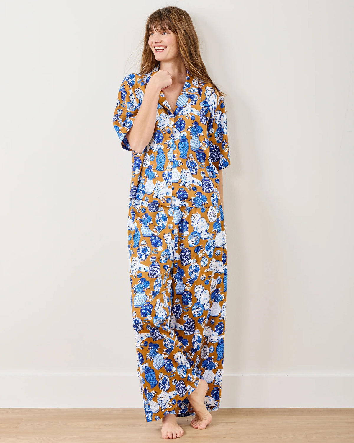 A person in Printfresh&#39;s Satin Fancy Cat Wildest Dreams pajama set, crafted from eco-friendly viscose and adorned with blue, white, and brown hand screen-printed motifs, stands barefoot by a plain wall. They&#39;re smiling with one hand touching their chin.