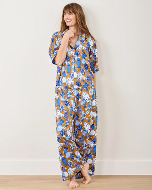 A person in Printfresh's Satin Fancy Cat Wildest Dreams pajama set, crafted from eco-friendly viscose and adorned with blue, white, and brown hand screen-printed motifs, stands barefoot by a plain wall. They're smiling with one hand touching their chin.