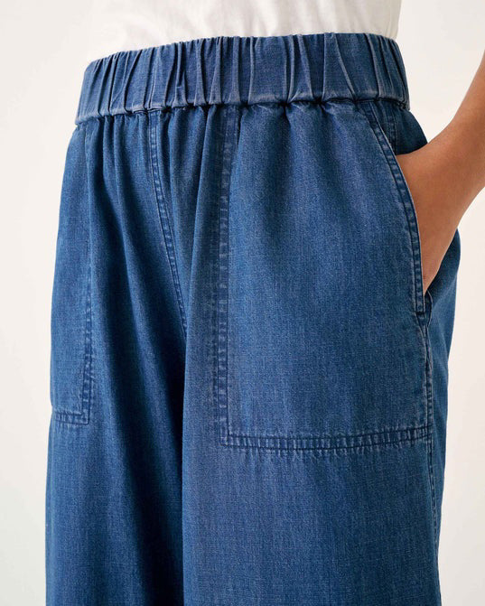 Wearing MERSEA - SAMMIE WIDE LEG PANT in light blue chambray with a pull-on waistband and front patch pockets, styled with a light top and cream sneakers, the person stands against a plain background.