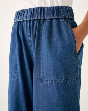 Wearing MERSEA - SAMMIE WIDE LEG PANT in light blue chambray with a pull-on waistband and front patch pockets, styled with a light top and cream sneakers, the person stands against a plain background.