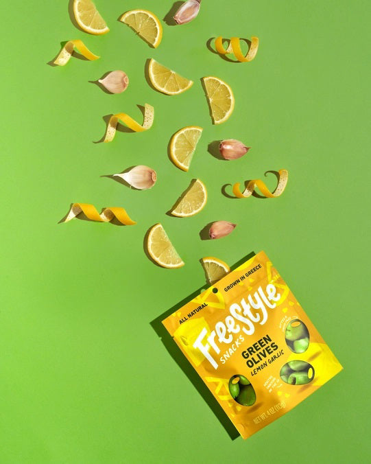 A yellow packet of FREESTYLE - LEMON GARLIC GREEN OLIVES from FREESTYLE SNACKS, featuring green olives flavored with extra virgin olive oil. The packaging highlights the "all-natural" ingredients and notes that the olives are "grown in Greece." Weighing 4 oz (113g), the pack showcases images of green olives alongside phrases such as "pitted &amp; unpasteurized" and "healthy snacking on the go.