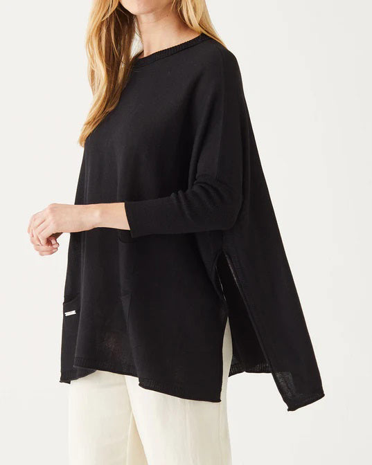 A smiling woman with long blonde hair wears the MERSEA Catalina Crewneck Sweater in black, featuring signature split sides, paired with light pants and standing against a plain white background.