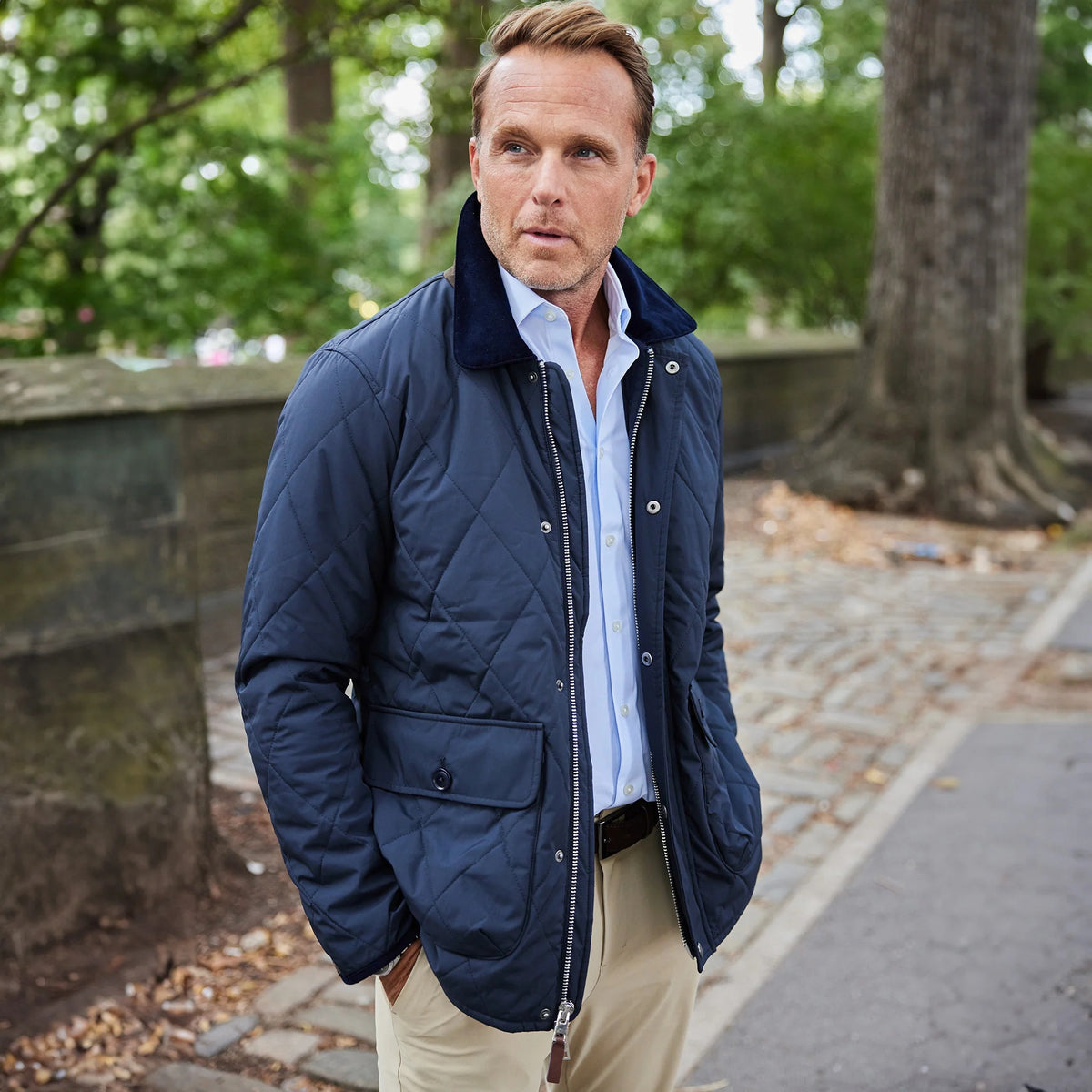 A man with light brown hair smiles while standing outside in a park. He wears the COLLARS AND CO - MIDDLEBURG QUILTED FIELD JACKET by COLLARS AND CO, featuring a corduroy collar, layered over a light blue shirt and tan pants for lightweight warmth on a cool day. Trees and a stone wall are visible in the background.