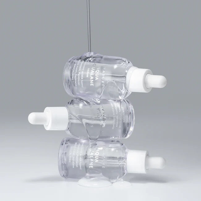 Three clear AQUA SQUALANE SERUM 50ML dropper bottles from ASIAN BEAUTY WHOLESALE are stacked with liquid dripping over them, set against a light gray background, showcasing this lightweight serum that helps tighten pores.