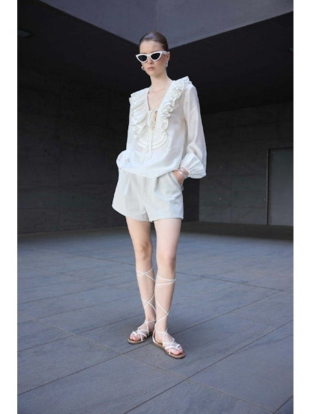A woman wearing white cat-eye sunglasses, the ELEH - BLOUSE WITH LACES AND RUFFLES by ELEH, and white shorts stands in a modern outdoor setting with grey walls and flooring.