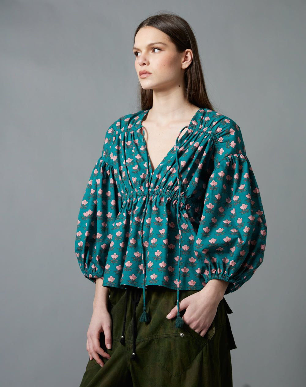 A person with long brown hair, seen from behind, wears the HOPE FOR FLOWERS Shirred Blouse and loose, dark green cargo pants with large pockets, standing against a plain gray background.