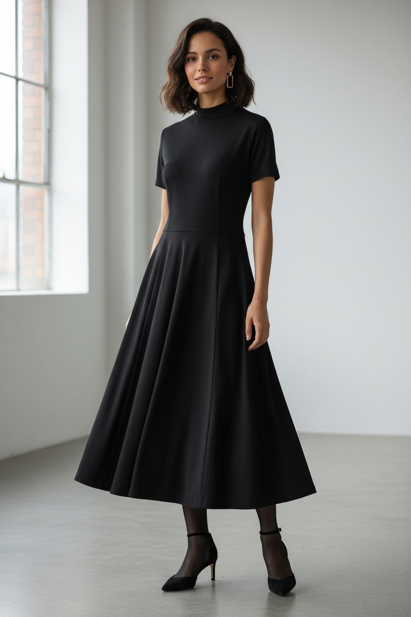 A woman faces away, wearing the LAVI COUTURE - MIDI DEIOPEA DRESS by LAVI in brown. The minimalist design features short sleeves and a fitted waist, styled with a neat bun and heeled sandals against a plain gray background.