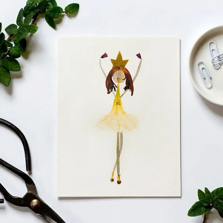 The PETAL PEOPLE PRESS - BALLERINA CARD by PETAL PEOPLE PRESS features a girl in a yellow dress and star headpiece, perfect as a thank you note card. Shown on a white surface with green leaves, scissors, and paperclips.