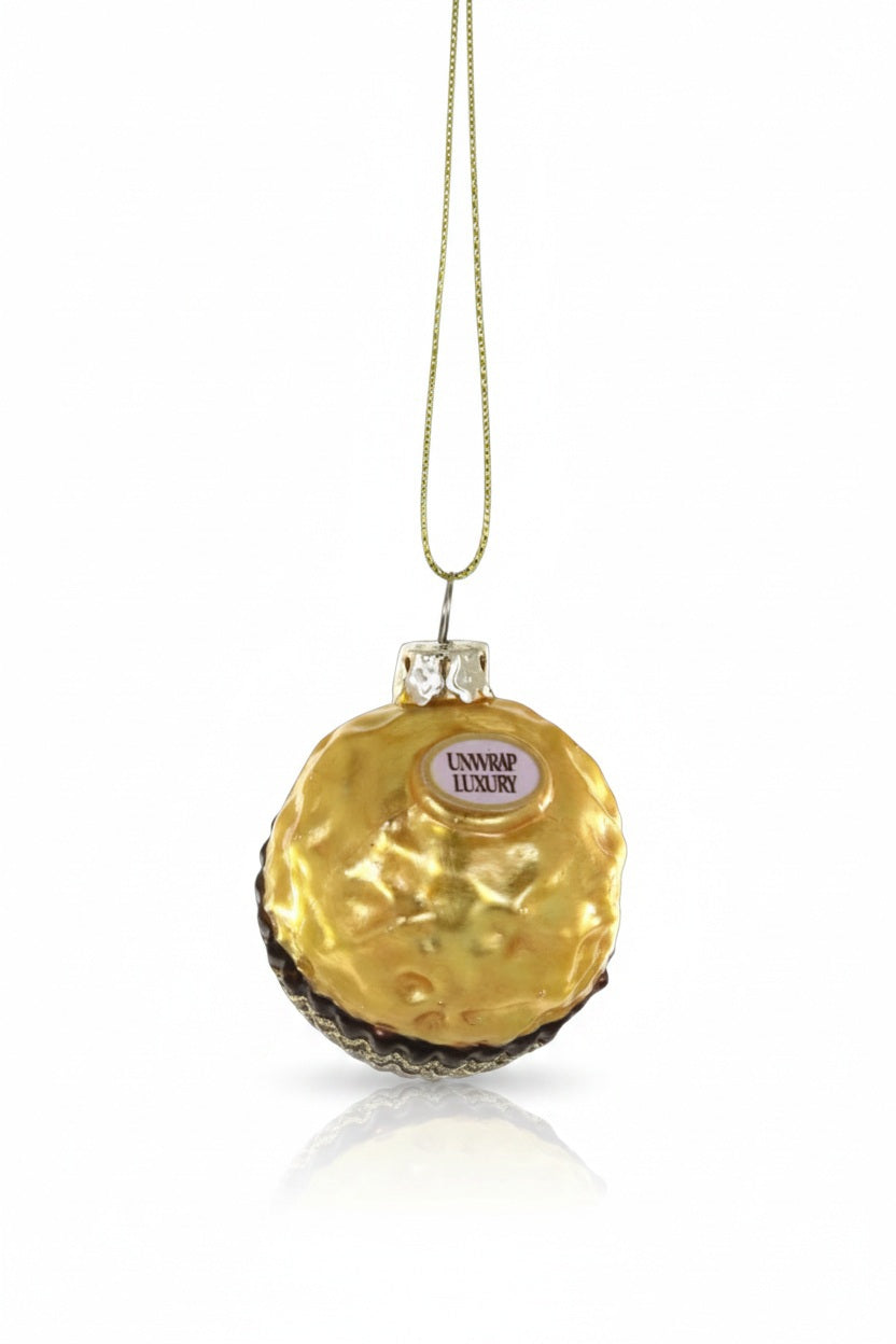 The CHOCOLATE HAZELNUT BON BON ORNAMENT by CODY FOSTER is a glass Christmas tree ornament shaped like a gold foil-wrapped candy with a "UNWRAP LUXURY" label, hanging by a gold string.