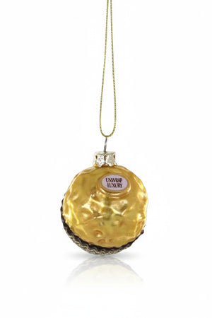 The CHOCOLATE HAZELNUT BON BON ORNAMENT by CODY FOSTER is a glass Christmas tree ornament shaped like a gold foil-wrapped candy with a "UNWRAP LUXURY" label, hanging by a gold string.