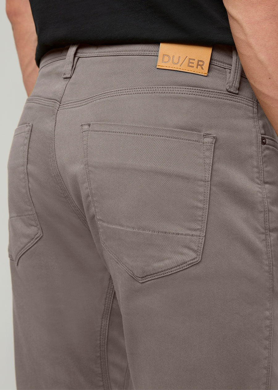Close-up of the back of a person wearing DUER No Sweat Athletic Taper Pants in Stone, featuring two back pockets and a tan leather DUER patch on the waistband. They're also wearing a black shirt.