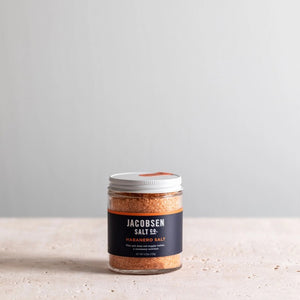 A small jar of JACOBSEN SALT CO. JACOBSEN SALT - INFUSED HABANERO SALT with a white lid sits on a light-colored surface against a plain, light gray background, ideal as spicy salt or for rimming cocktails.