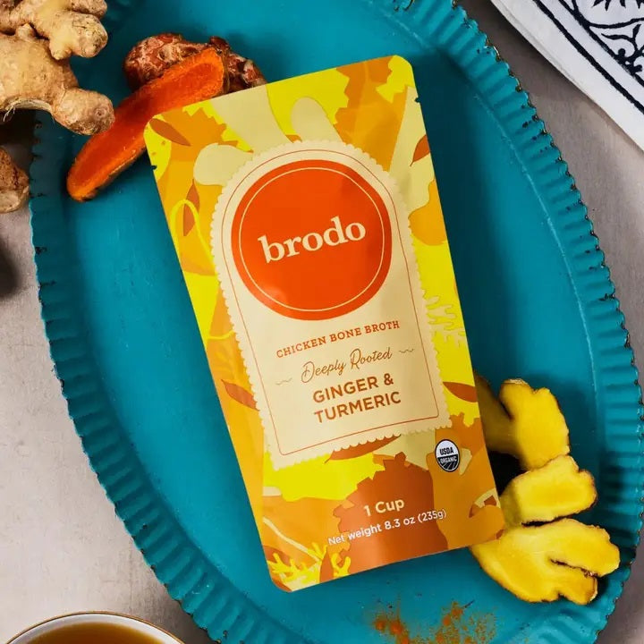 A carton of BRODO BROTH- DEEPLY ROOTED TURMERIC & GINGER BONE BROTH ORGANIC by BRODO BROTH COMPANY rests on a blue tray, surrounded by fresh turmeric and ginger roots—an ideal choice for those seeking a keto-friendly option.
