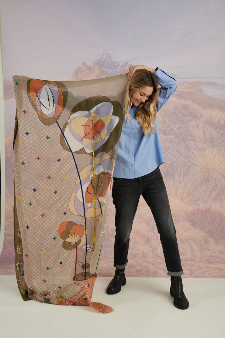 A woman with long blonde hair, in a light blue shirt and dark pants, smiles in front of a scenic backdrop, draping the elegant, hand-embroidered STORIATIPIC - ASTER WOOL SCARF around her shoulders.