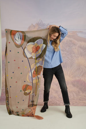 A woman with long blonde hair, in a light blue shirt and dark pants, smiles in front of a scenic backdrop, draping the elegant, hand-embroidered STORIATIPIC - ASTER WOOL SCARF around her shoulders.