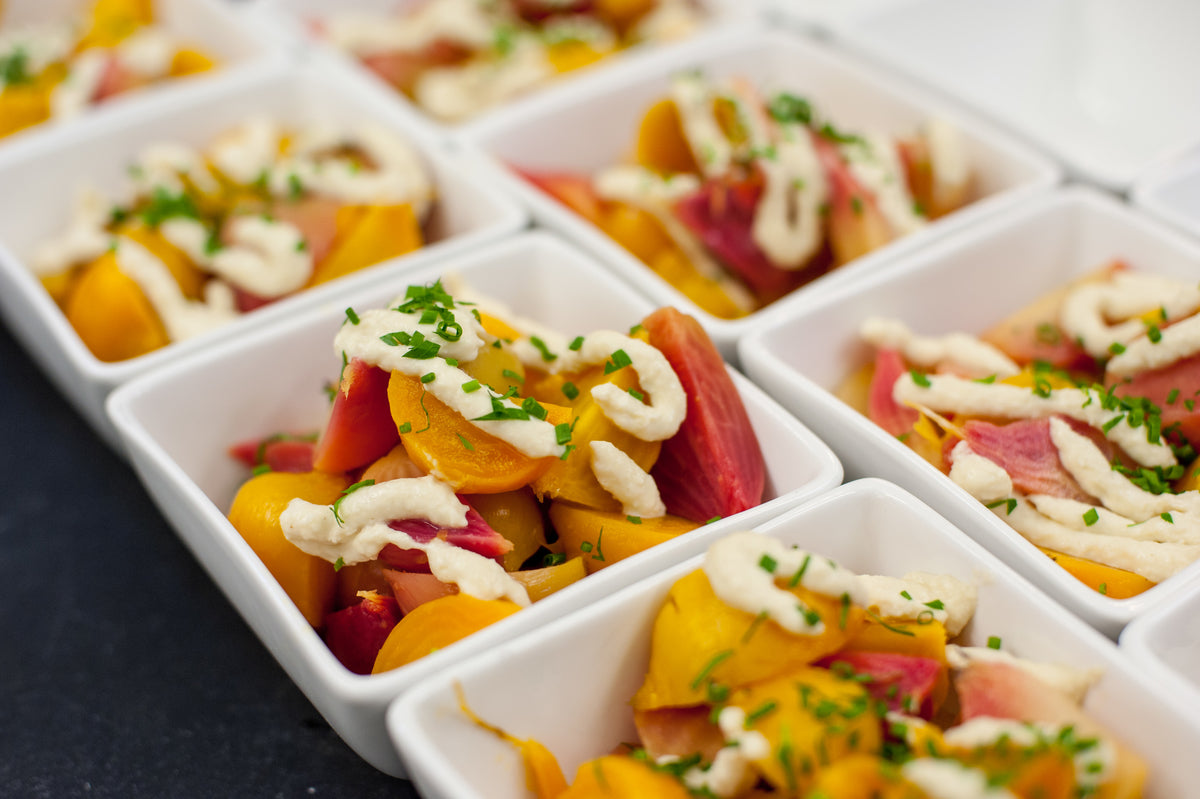 Small white bowls filled with colorful roasted beets, topped with a swirl of creamy white sauce and garnished with chopped fresh herbs, arranged in neat rows on a dark surface.