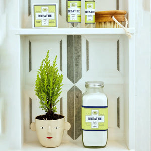 A small green plant in a face-shaped pot sits next to a large jar labeled "BAUDELAIRE - BREATHE SHOWER MIST." Above, smaller bottles of eucalyptus essential oil and a wooden brush rest on a white shelf with a gray geometric background, creating an inviting space for your aromatherapy steam room.