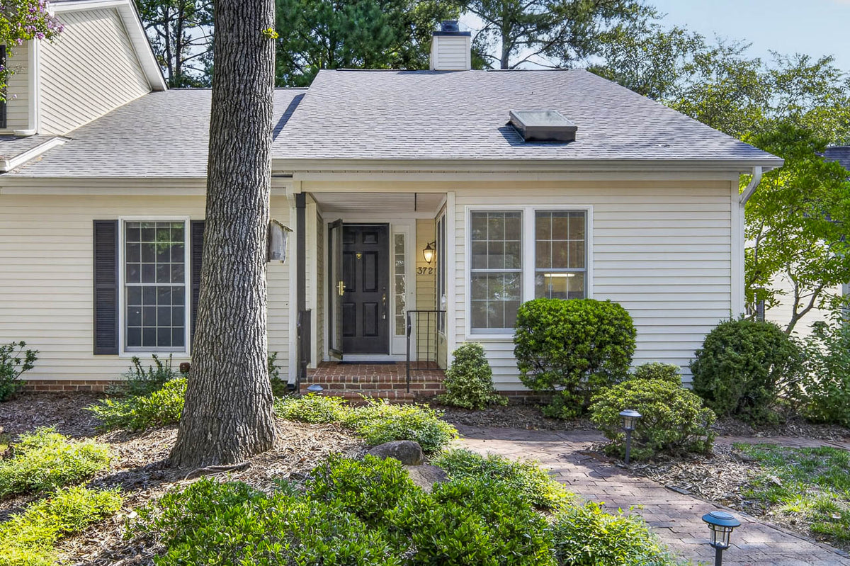 A small house with cream siding, black shutters, and a black front door. There are bushes, a tree, and a brick walkway leading to the entrance, with a porch light next to the door.