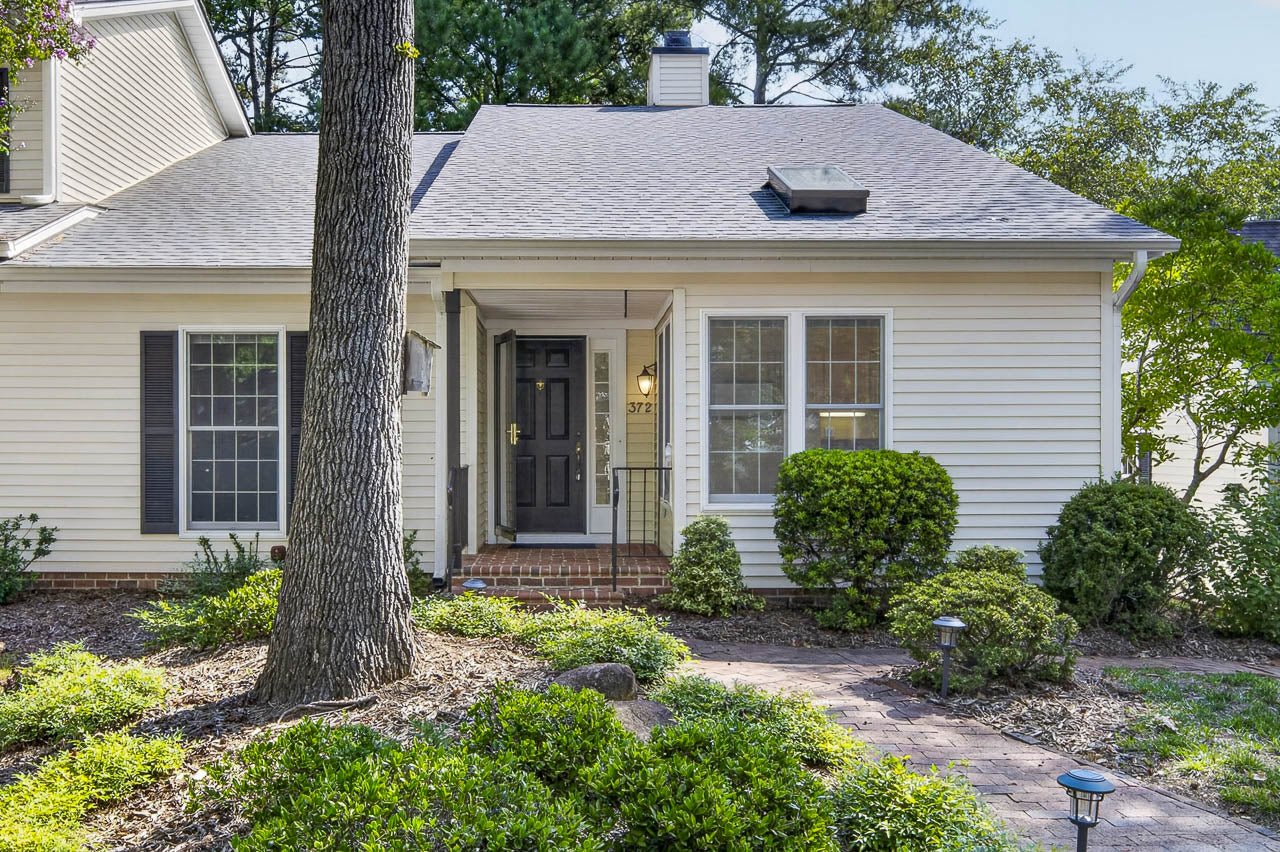 Single-story beige house with black shutters, a black front door, and a small covered porch. Brick steps lead to the entrance, surrounded by shrubs and a garden path with mulch and greenery.