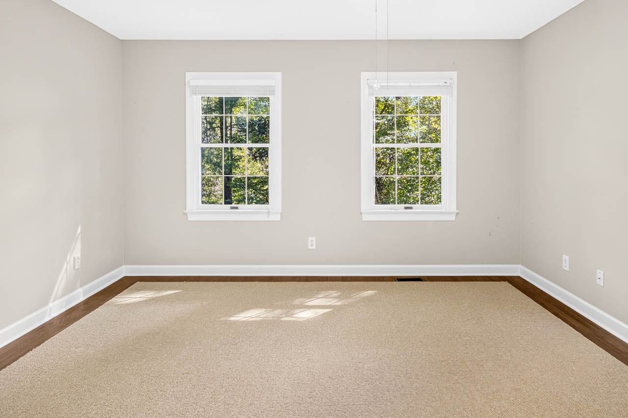 Empty room with beige carpet, light tan walls, white trim, and two windows letting in natural light. The windows show green trees outside. The floor has a border of dark wood around the carpet.