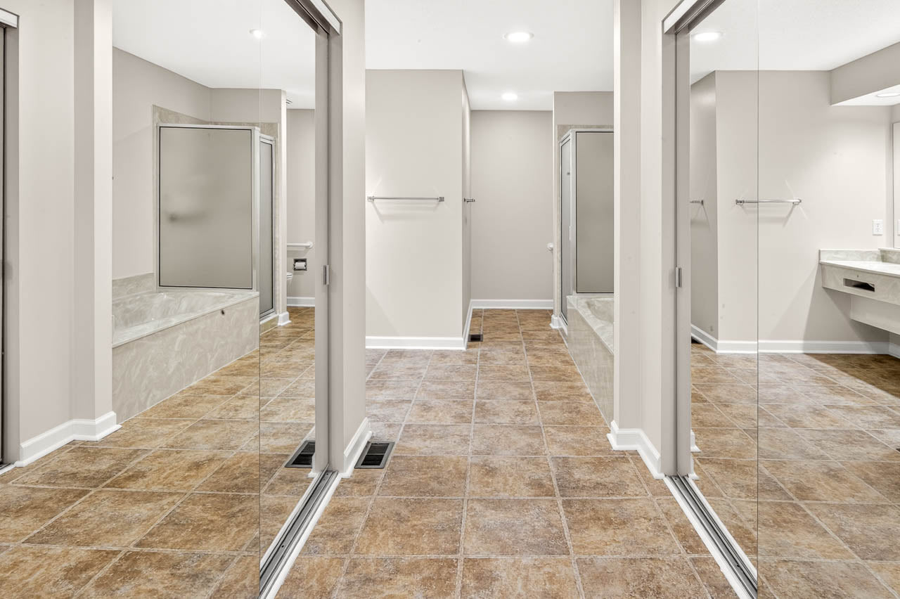 Modern bathroom with brown tile floor, beige walls, mirrored closet doors, two bathtubs with glass screens, towel racks, and a vanity area with a sink and countertop on the right.