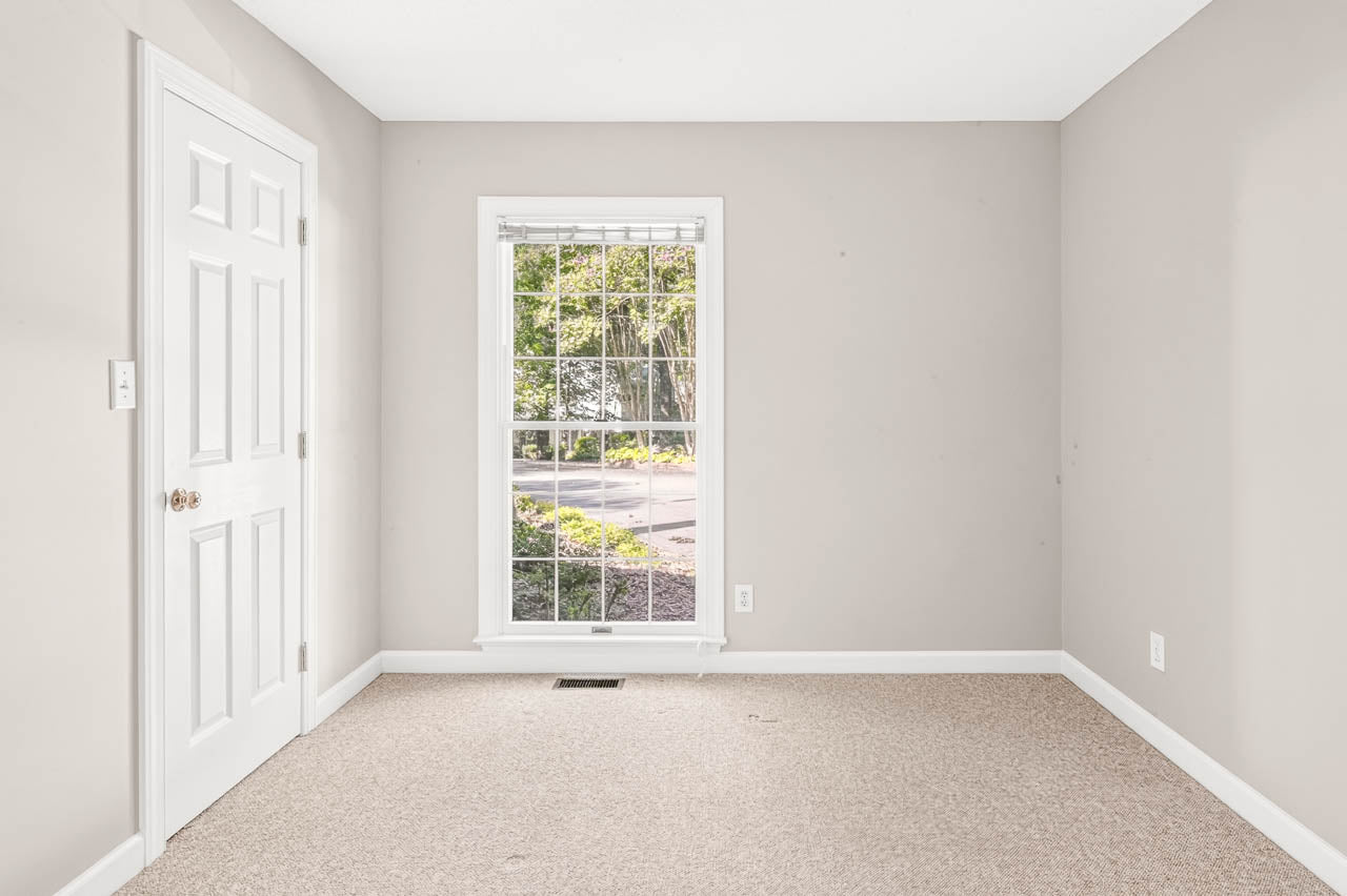 Empty room with beige carpet, light gray walls, a white door with two knobs on the left, and a large window with white framing overlooking greenery and a road outside.