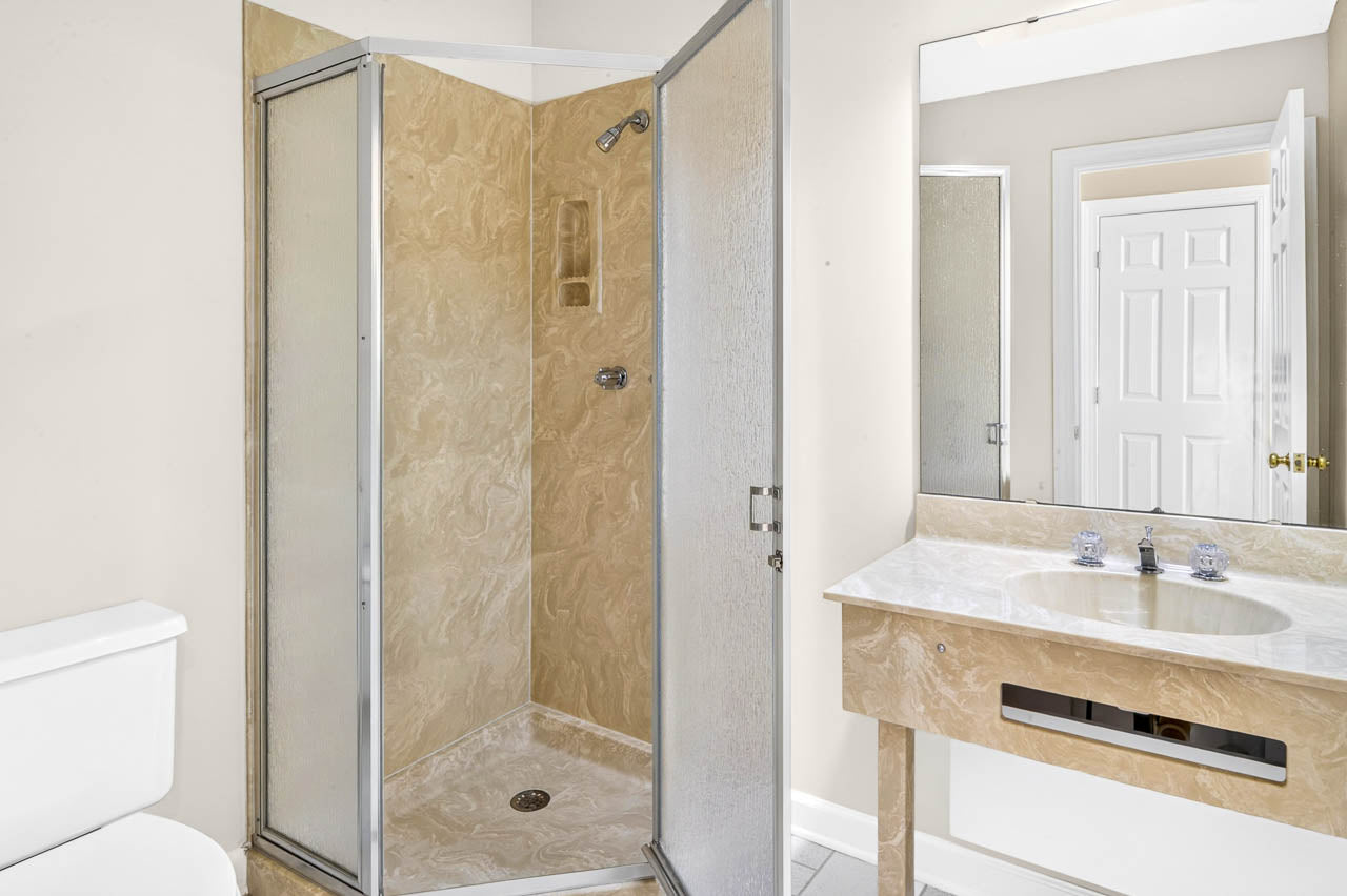 A bathroom with a beige shower stall, glass door, white toilet, and a marble-patterned sink vanity with a large mirror above it. A closed white door is visible in the background.