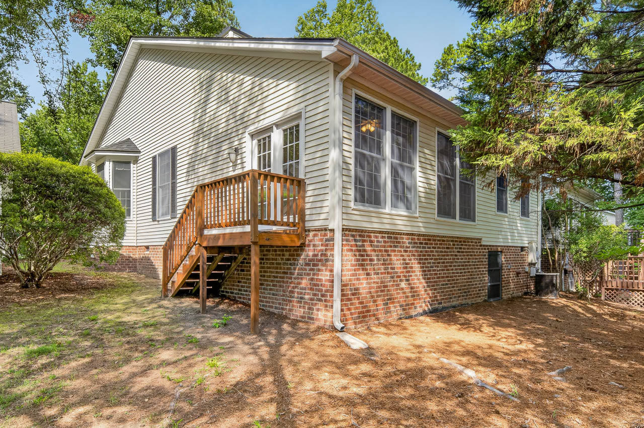Single-story house with beige siding, brick foundation, and black shutters. A small wooden deck with stairs leads to a glass door. Mature trees and bushes surround the home, and the yard is covered with pine straw.