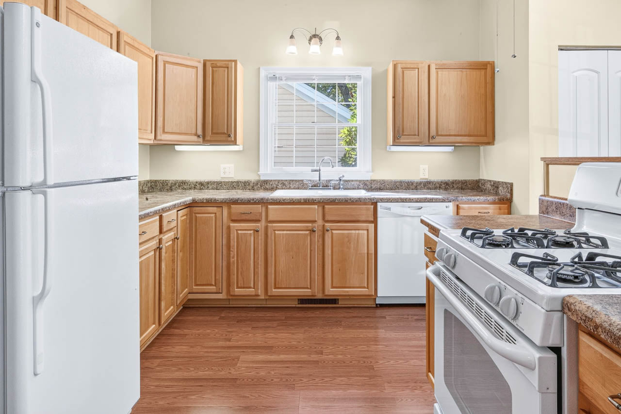 Kitchen with light wood cabinets, white refrigerator, dishwasher, and gas stove, brown countertops, double sink under a window with blinds, and overhead light fixture, all on wood-style flooring.